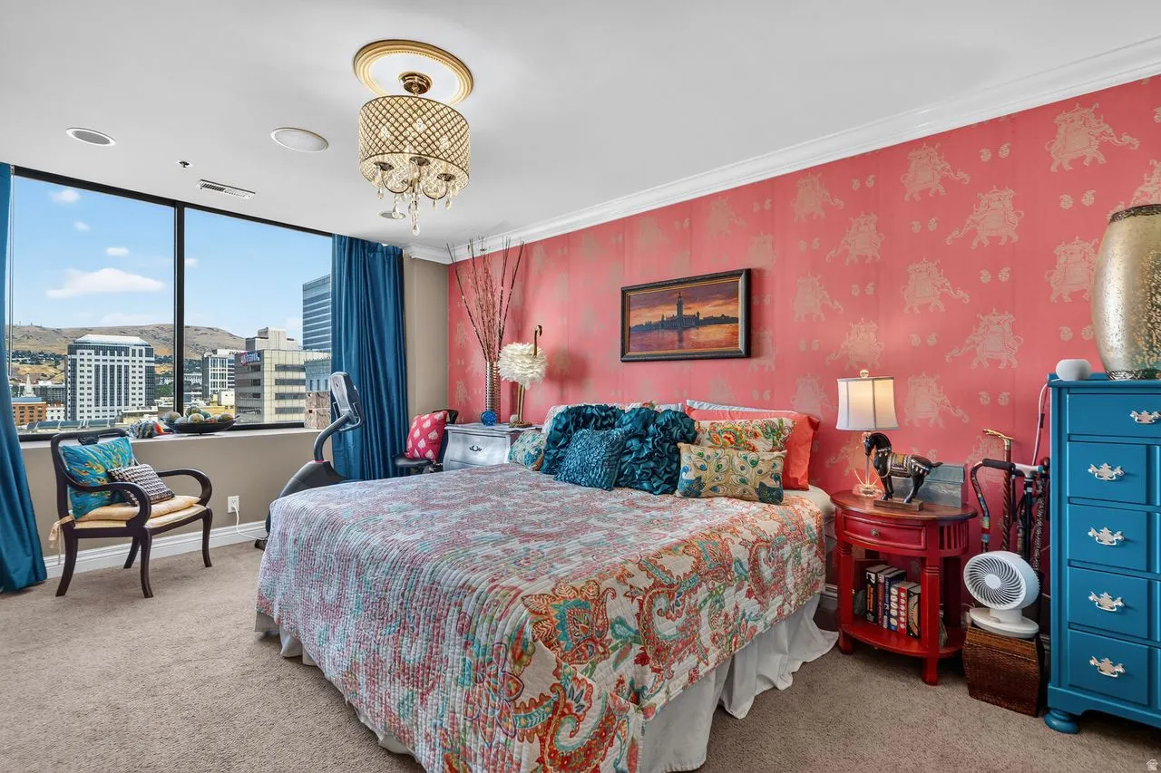 Sophisticated bedroom retreat featuring floor-to-ceiling windows with panoramic city skyline views, vibrant coral damask wallcovering, plush carpeting, brass ceiling fixture, and blue painted built-in cabinetry with multiple drawers