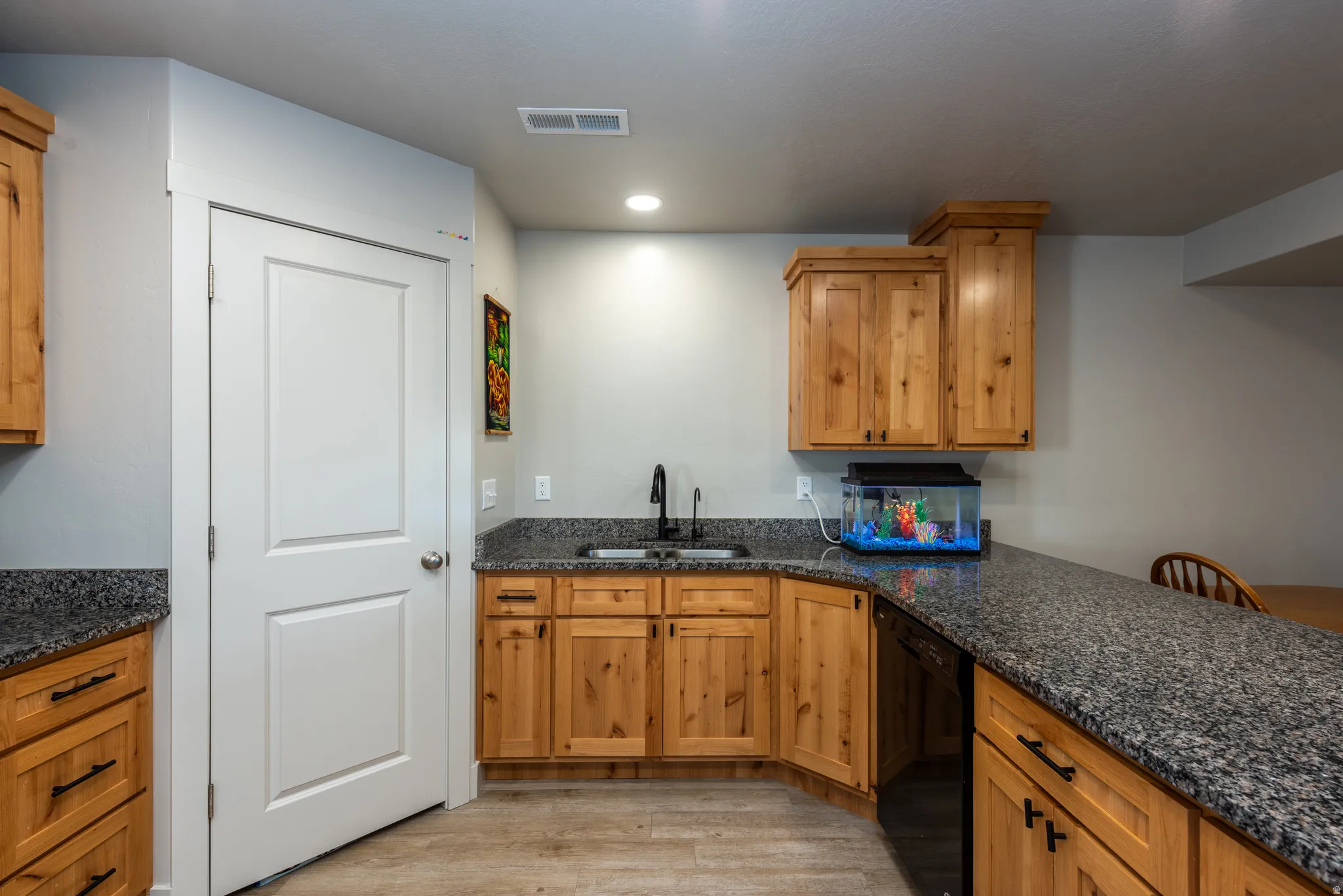 Functional kitchenette featuring granite-look countertops, a stainless steel sink with a black faucet, natural wood cabinetry, and wood-finish flooring