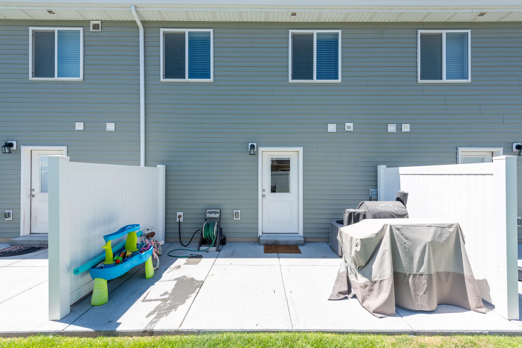 Modern home exterior featuring a spacious concrete patio with direct access to the residence, vinyl horizontal siding in a soft blue-gray finish, multiple double-hung windows with dark frames, and a manicured lawn