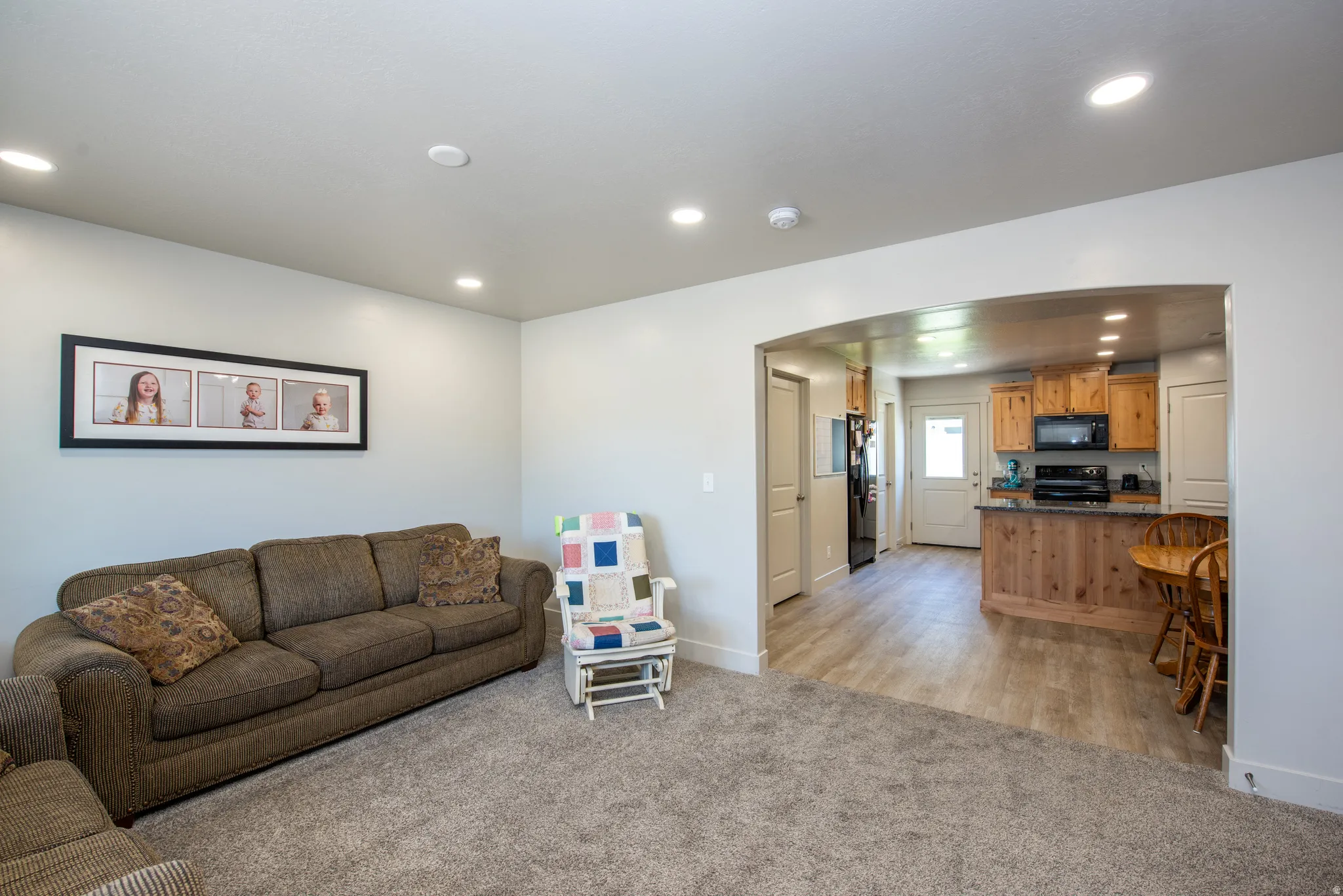 Carpeted living area featuring recessed lighting, an arched entryway, and direct access to a kitchen with wood cabinetry, a granite-look countertop, and wood-finish flooring
