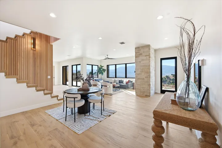 Open-concept interior featuring light wood-finish flooring, a wood-paneled accent wall with integrated staircase, a stone-clad architectural pillar, and multiple floor-to-ceiling windows with expansive mountain scenery