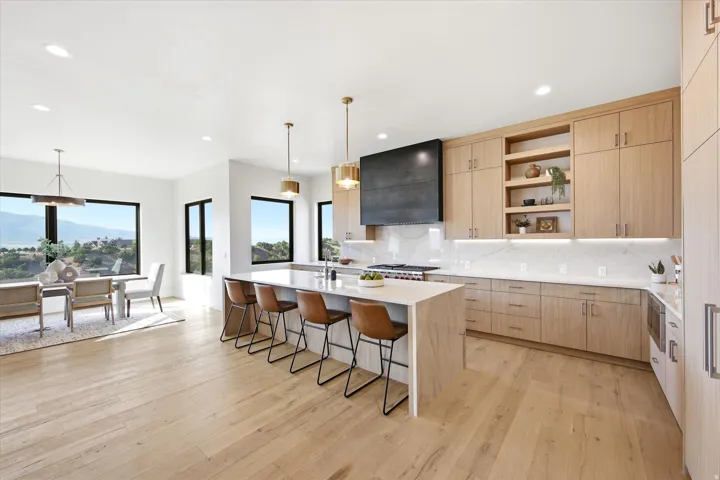 Open-concept kitchen with light wood-finish flooring, a large island with seating, extensive cabinetry with integrated shelving, a range with a black range hood, and a dining area with multiple windows