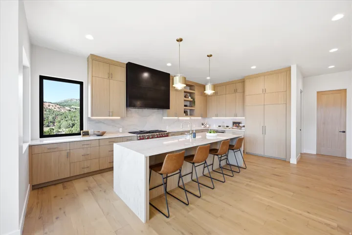 Contemporary kitchen featuring custom wood-finish cabinetry, a white quartz-topped island with seating for multiple guests, professional-grade gas cooktop, stainless steel appliances, pendant lighting, and wide-plank wood-finish flooring throughout
