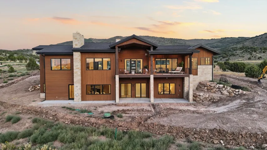 Contemporary architecture featuring wood-finish siding, stone accents, multiple large windows, a spacious upper-level balcony, and a ground-level patio