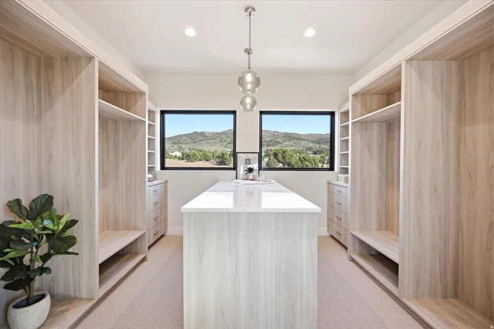 Integrated custom closet system with light wood-finish cabinetry, central island with a light stone countertop, recessed lighting, and a contemporary pendant fixture