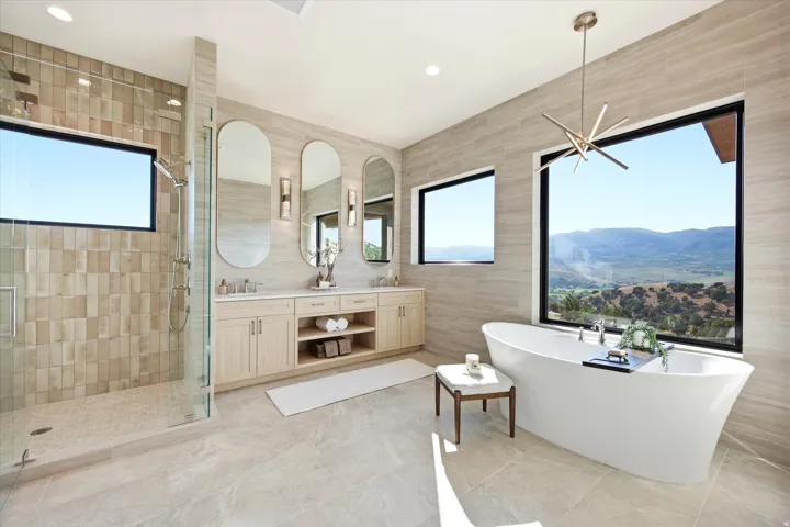 Contemporary bathroom featuring a freestanding soaking tub, expansive window with mountain scenery, dual vanity with oval mirrors, glass-enclosed shower, and large format tile flooring