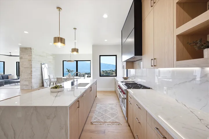 Modern kitchen featuring a waterfall island, light wood-finish cabinetry, integrated stainless steel appliances, and large black-framed windows showcasing distant scenery