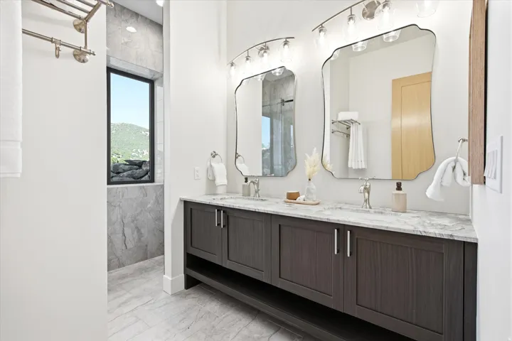 Spacious bathroom featuring a dual vanity with a light-toned countertop, two decorative mirrors with overhead lighting, a large window, and light-toned tile flooring