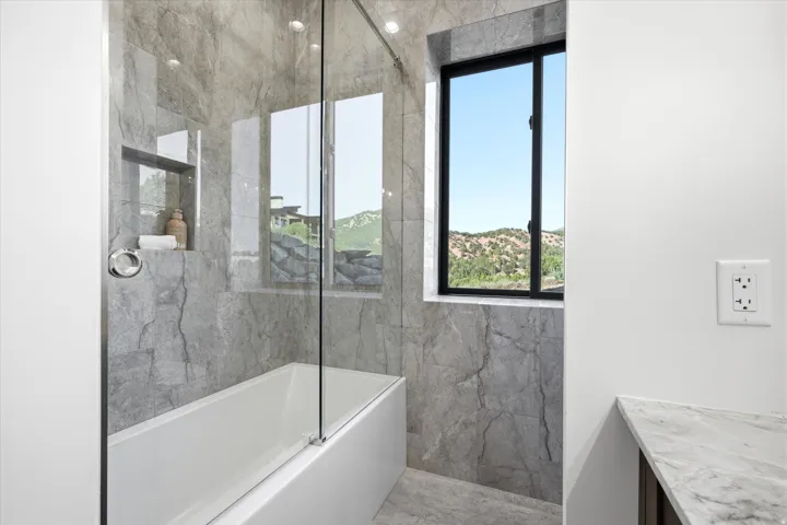 Bathtub with full-height tile surround, glass shower enclosure, integrated shower niche, and a window with a mountain backdrop