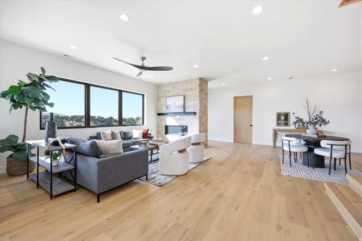 Expansive open-concept living area featuring light wood-finish flooring, a stone-clad fireplace with a dark mantel, large multi-pane windows, and recessed lighting
