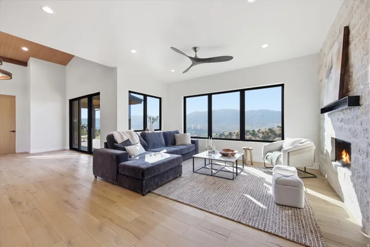 Spacious living area featuring wood-finish flooring, a textured stone-clad fireplace with mantel, and large picture windows showcasing mountain scenery