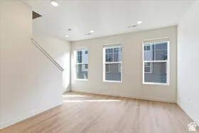 Bright open-concept interior featuring light wood-finish flooring, recessed ceiling lighting, three double-pane windows with roller shades, and a staircase with a minimalist railing