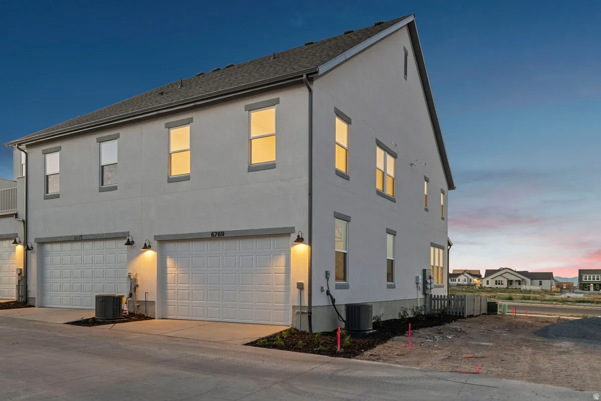 Modern townhome exterior featuring light-colored stucco siding, a dark shingled roof, multiple windows with dark trim, and two individual garage doors