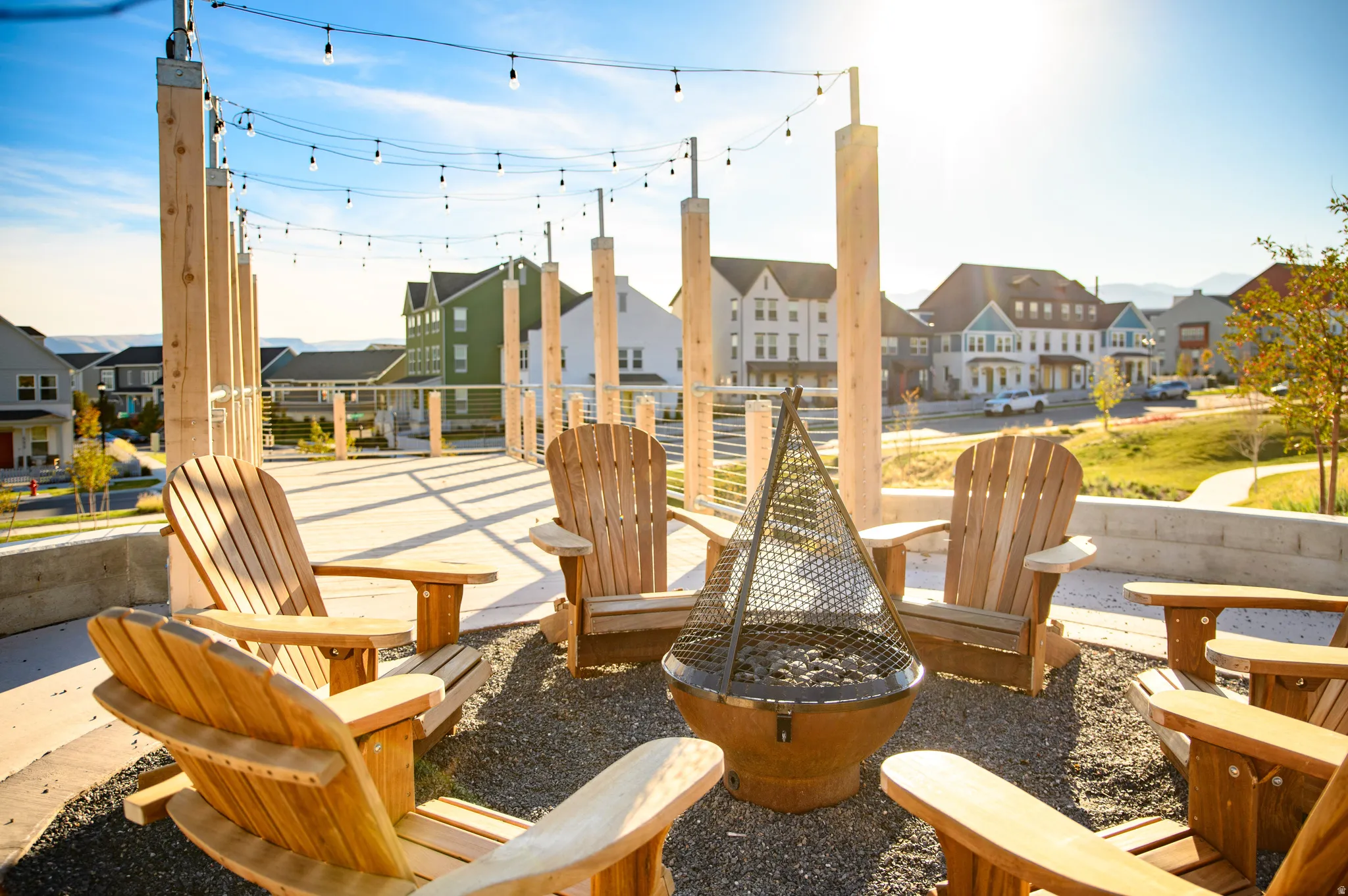 Outdoor amenity space featuring a gravel fire pit area with Adirondack chairs, overhead string lighting, a concrete retaining wall, and a paved pathway
