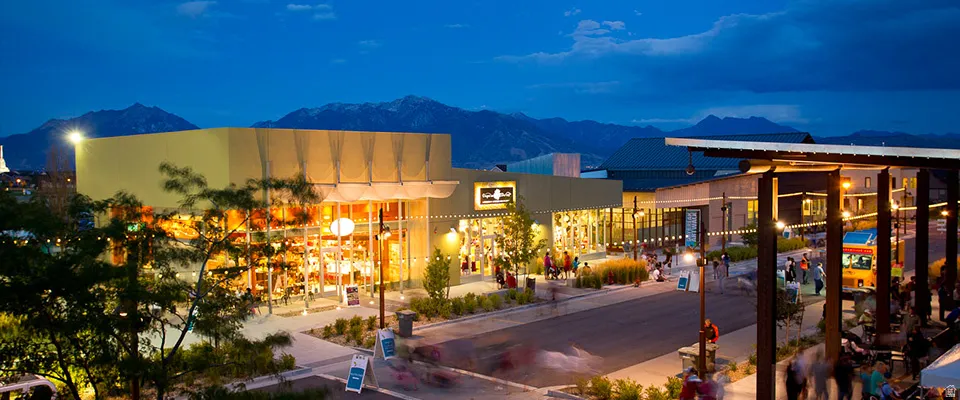 Modern commercial center featuring multiple storefronts with large display windows, contemporary architecture, pedestrian walkways, and mountain scenery