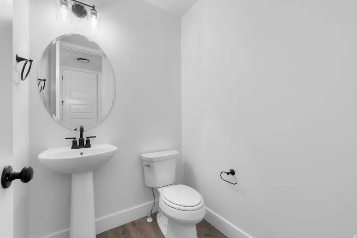 Pedestal sink, black-finish hardware, oval mirror, wall-mounted light fixture, and wood-finish flooring