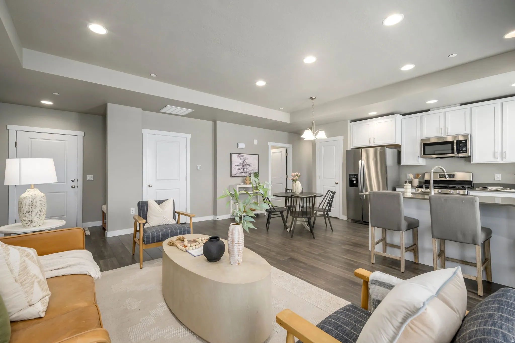 Open-concept living and dining area featuring recessed ceiling lighting, a curved upholstered ottoman, wood-finish flooring, and direct sightlines to a modern kitchen with stainless steel appliances, white cabinetry, and a quartz-topped island with seatin