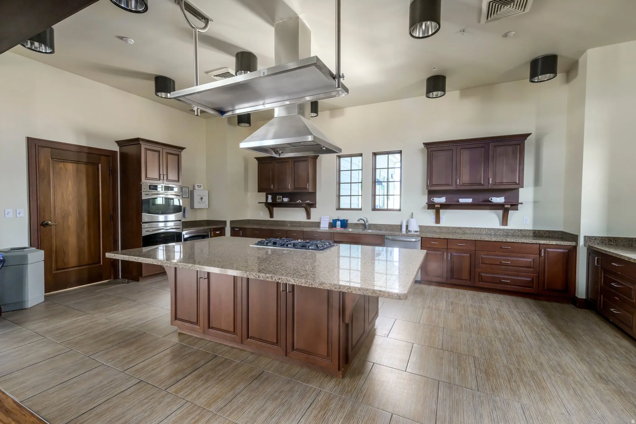 Spacious kitchen featuring an expansive center island with a built-in gas range, stainless steel range hood, stainless steel double wall oven, wood-finish cabinetry, and light-toned granite countertops