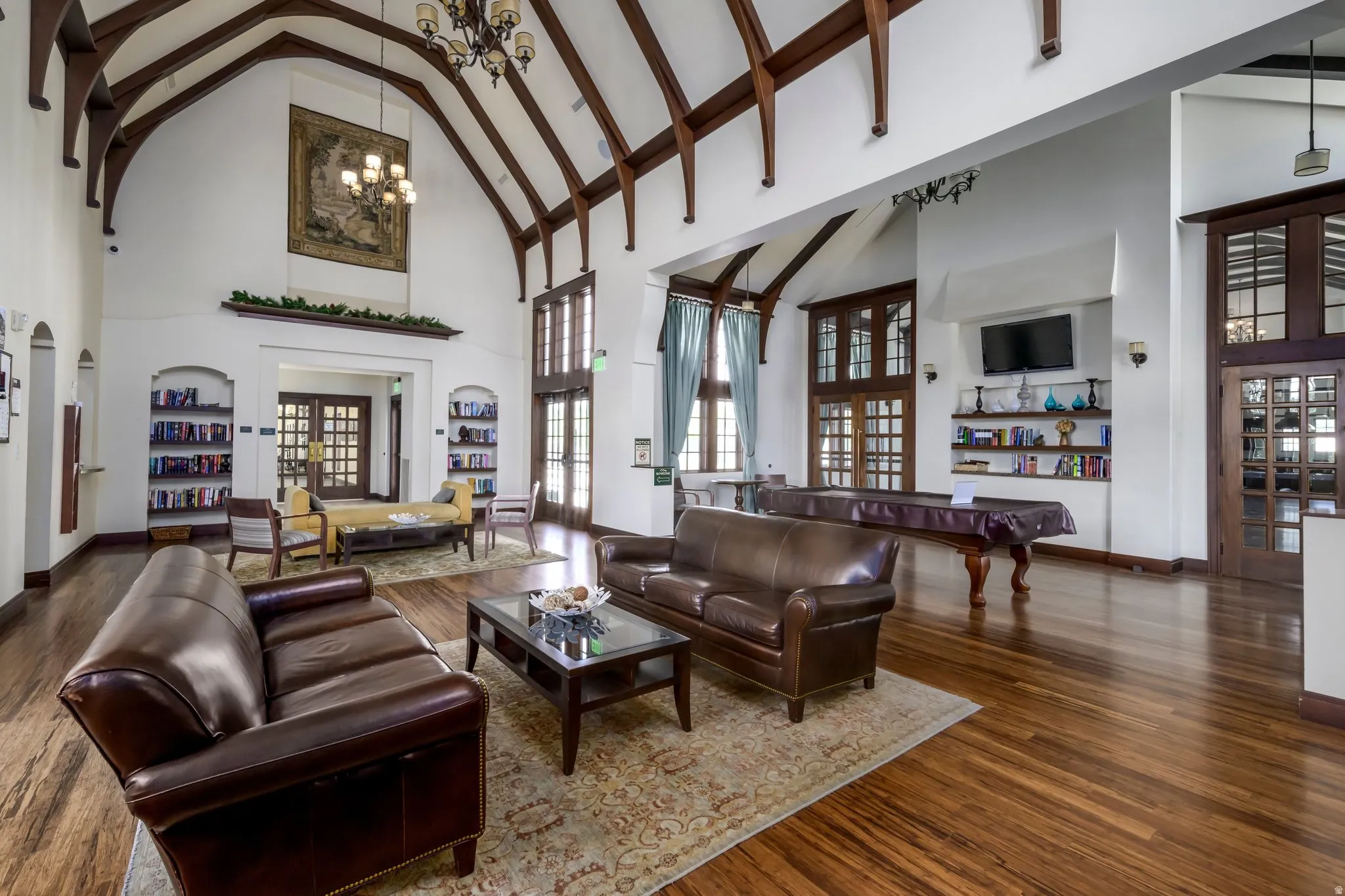 Spacious common area featuring vaulted ceilings with exposed wood beams, wood-finish flooring, built-in shelving, and multiple large windows with transom details