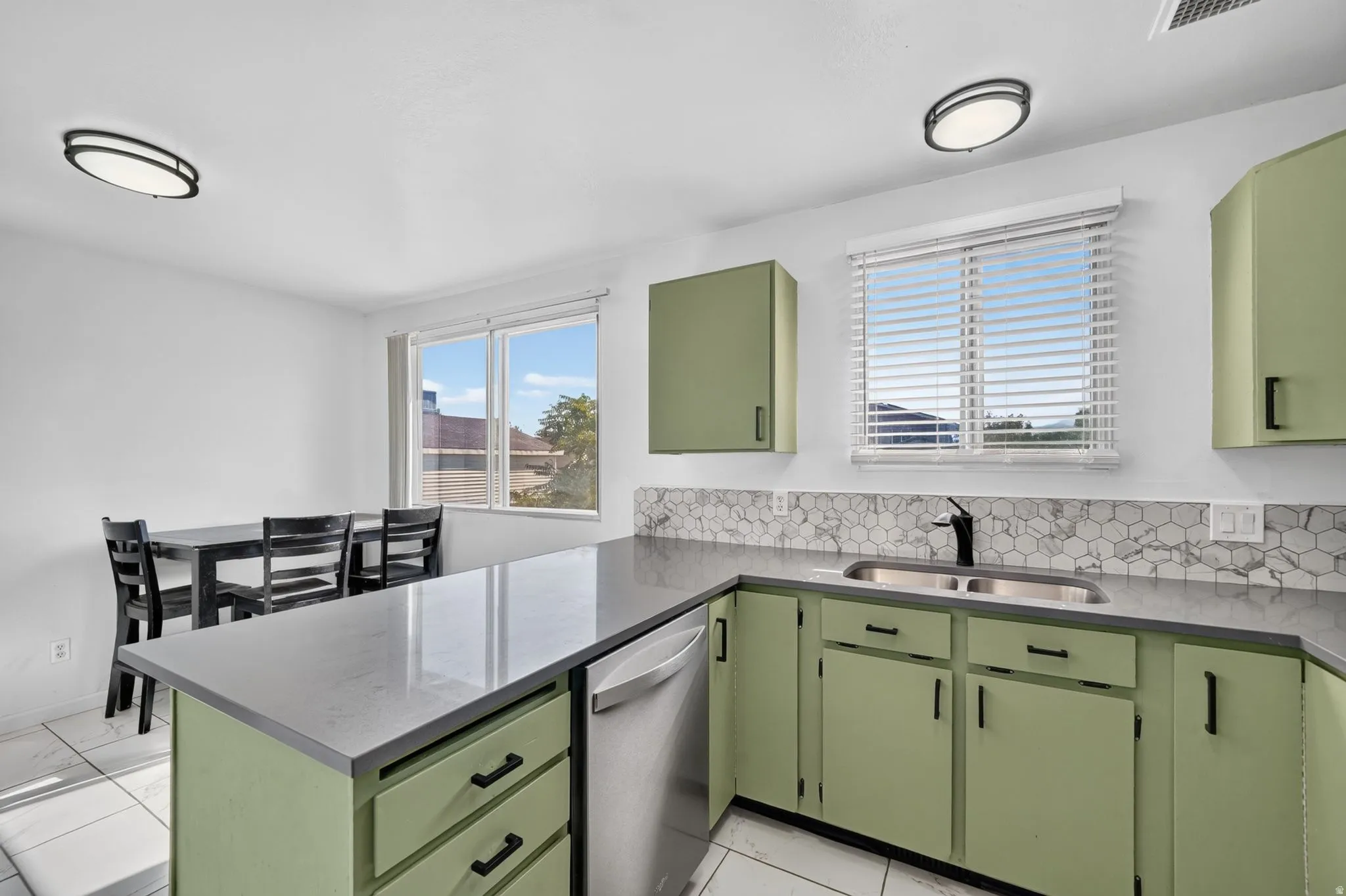 Kitchen featuring light green cabinetry, grey countertops, stainless steel dishwasher, and a hexagonal tile backsplash
