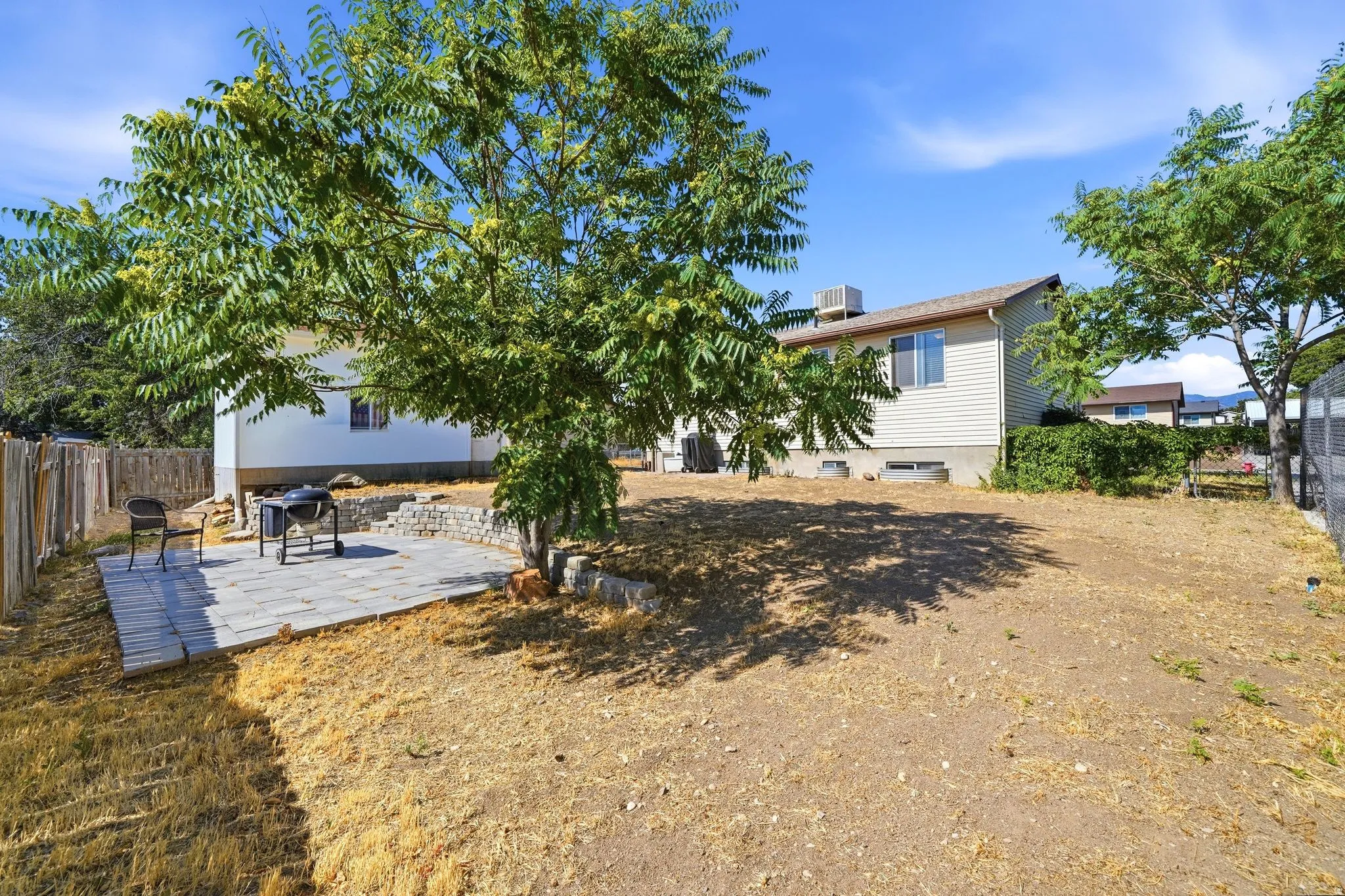 Expansive backyard featuring a stone paver patio, a wooden privacy fence, mature trees providing natural shade, and a visible portion of the property's siding exterior