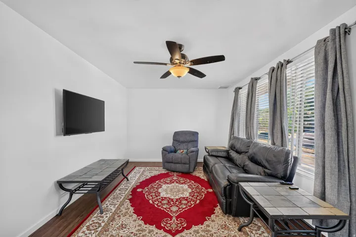 Living area featuring white walls, wood-finish flooring, a ceiling fan with integrated lighting, and multiple windows with horizontal blinds and drapery