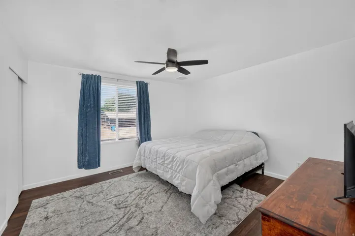 Bright bedroom featuring wood-finish flooring, a ceiling fan with integrated lighting, and a window with blinds and blue drapes