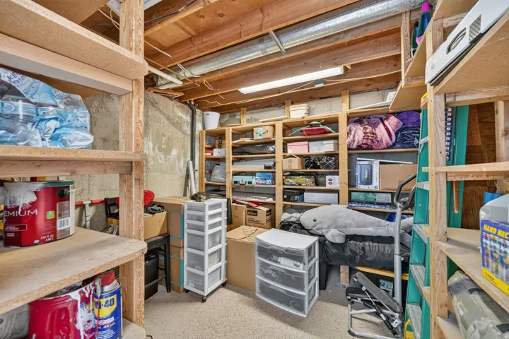 Basement storage area featuring extensive built-in wooden shelving, exposed ceiling joists, concrete walls, and concrete flooring