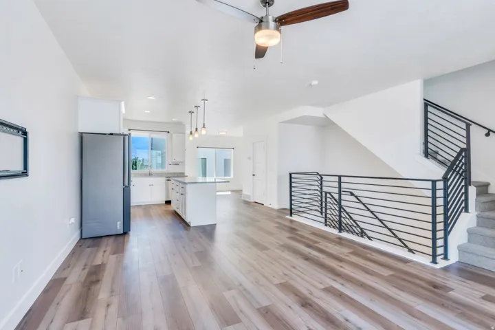 Open-concept living space featuring wood-finish flooring, a modern kitchen with white cabinetry and an island, a stainless steel refrigerator, and a staircase with contemporary black metal railings