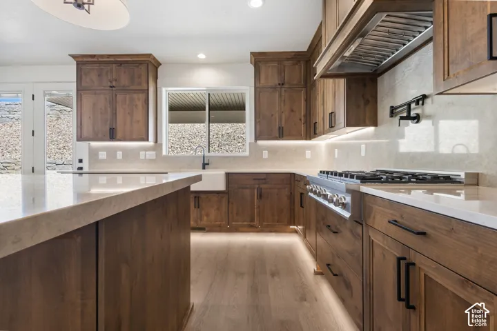 Kitchen featuring extensive wood cabinetry, light-toned countertops, integrated under-cabinet lighting, a built-in range with a professional hood vent, and wood-finish flooring