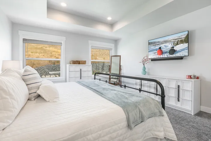Carpeted bedroom featuring a tray ceiling with recessed lighting, two windows with window coverings, a wall-mounted television, and a black metal bed frame