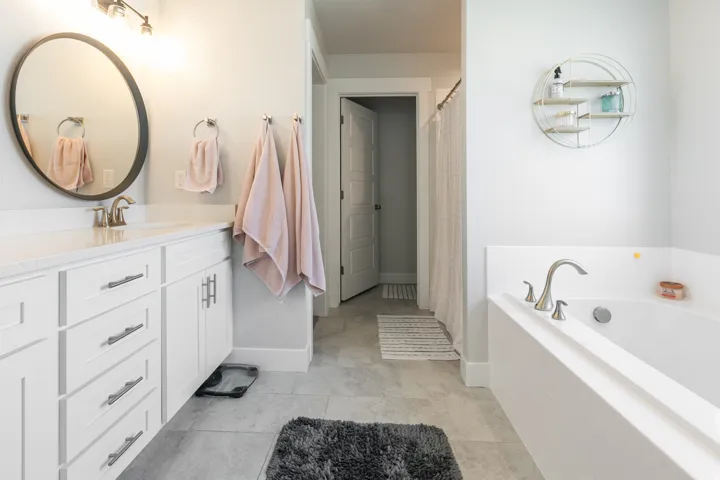 Bathroom featuring a white vanity with brushed nickel hardware, a circular mirror, an undermount sink, and a built-in bathtub with brushed nickel fixtures