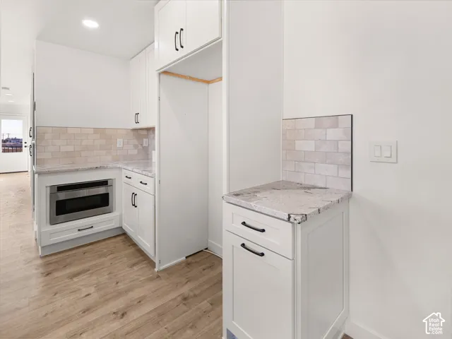 White cabinetry with black hardware, stone-finish countertops, tile backsplash, built-in microwave, and wood-finish flooring