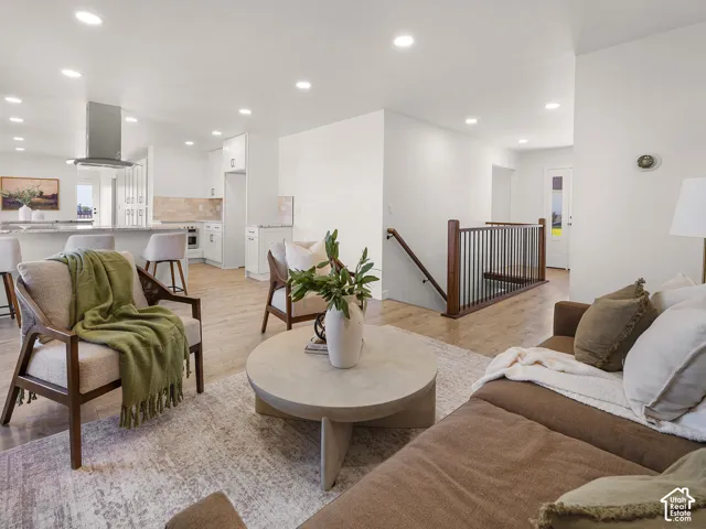 Open-concept living area featuring recessed lighting, wood-finish flooring, a kitchen island with seating, a stainless steel range hood, and a staircase with a dark wood and metal railing