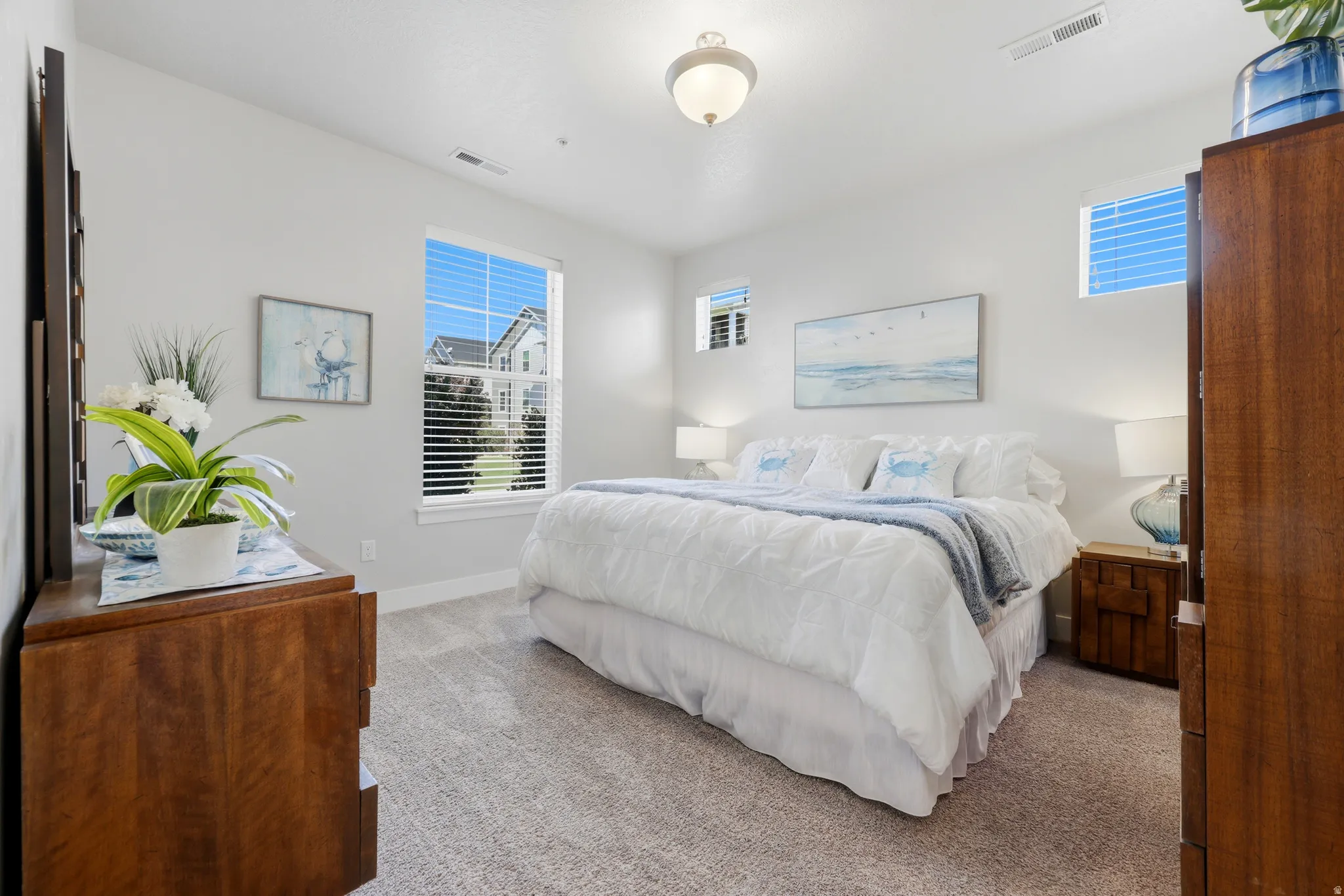 Serene primary bedroom showcasing abundant natural light from multiple windows, warm wood-finish furnishings, soft neutral bedding, plush carpeting, and a ceiling-mounted light fixture