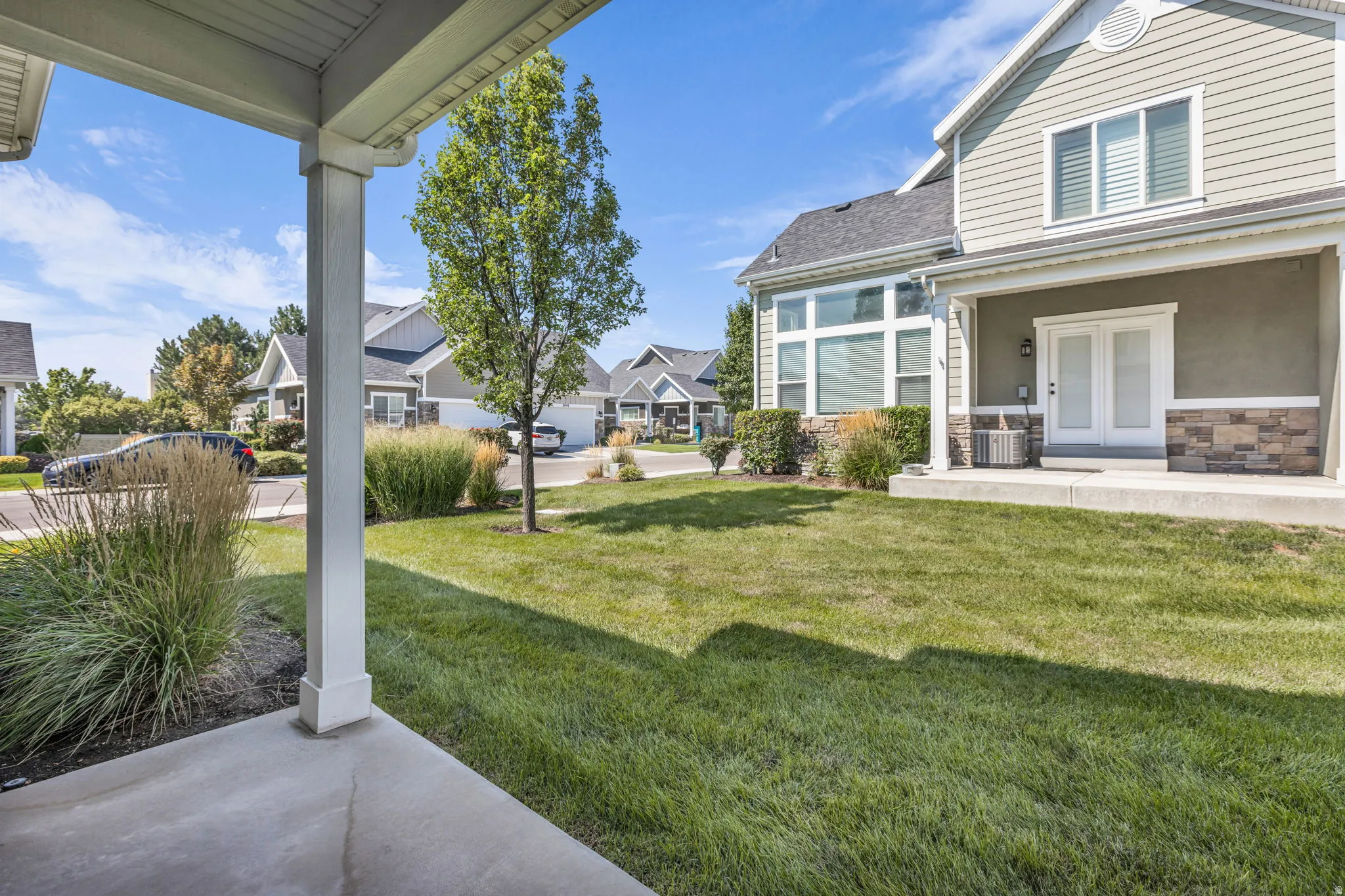 Covered patio entry with white pillars, green lawn, established tree, and decorative landscaping