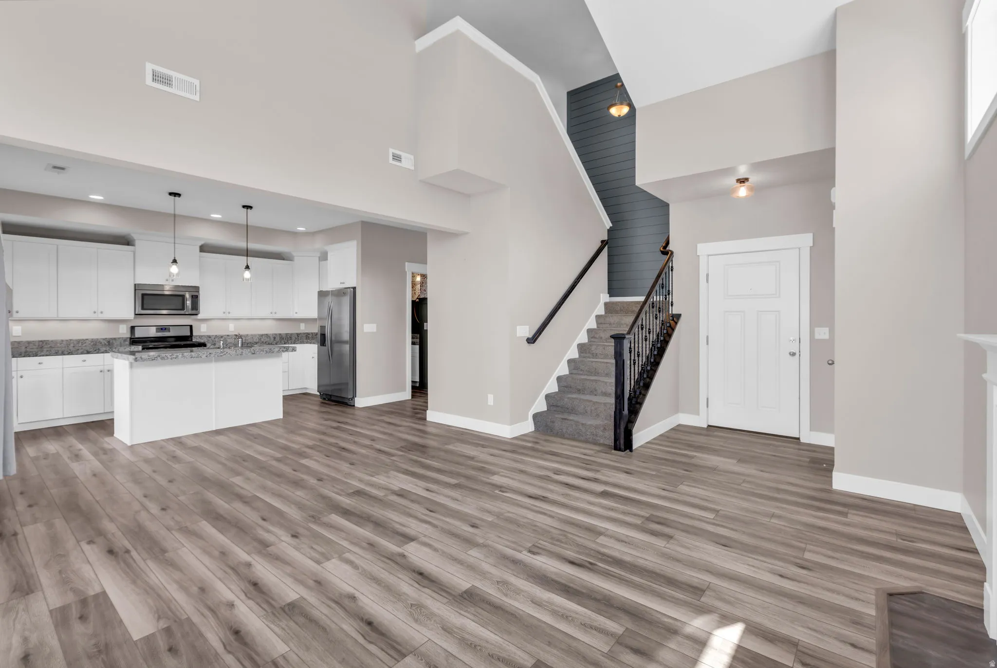 Open-concept living area featuring wood-finish flooring, a contemporary kitchen with white cabinetry, a kitchen island with seating, stainless steel appliances, and a staircase with a wrought iron baluster