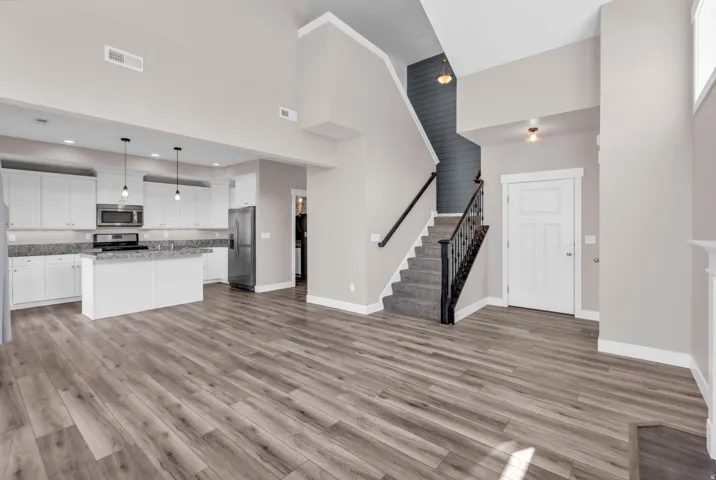 Open-concept living area featuring wood-finish flooring, a contemporary kitchen with white cabinetry, a kitchen island with seating, stainless steel appliances, and a staircase with a wrought iron baluster