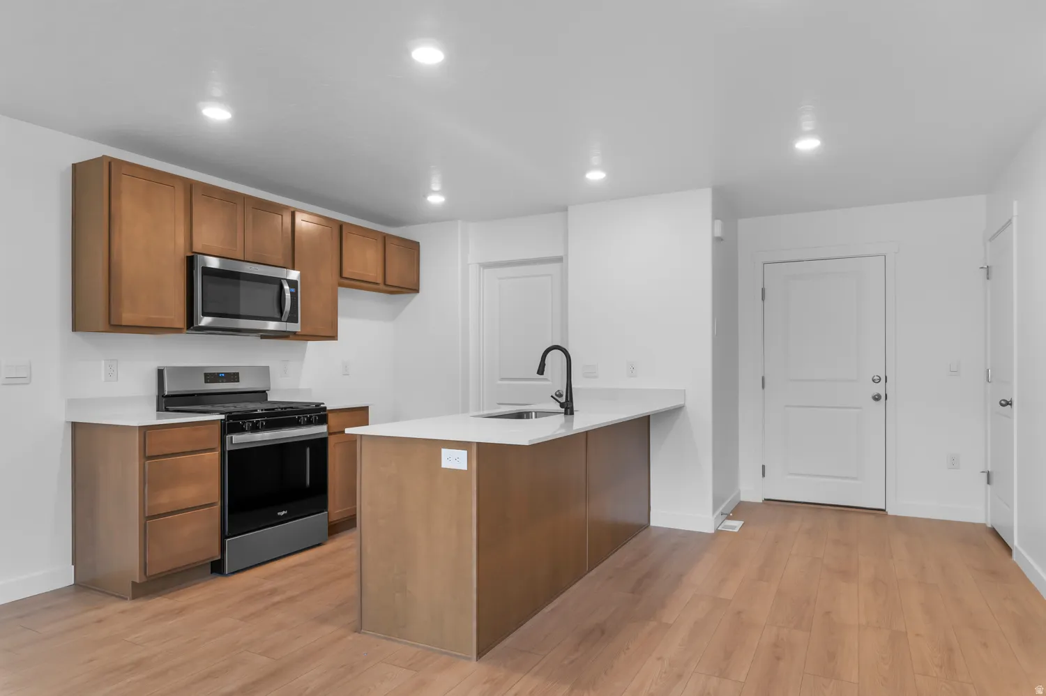 Open-concept kitchen featuring wood-finish cabinetry, stainless steel appliances, a kitchen island with a black faucet, white countertops, and recessed lighting