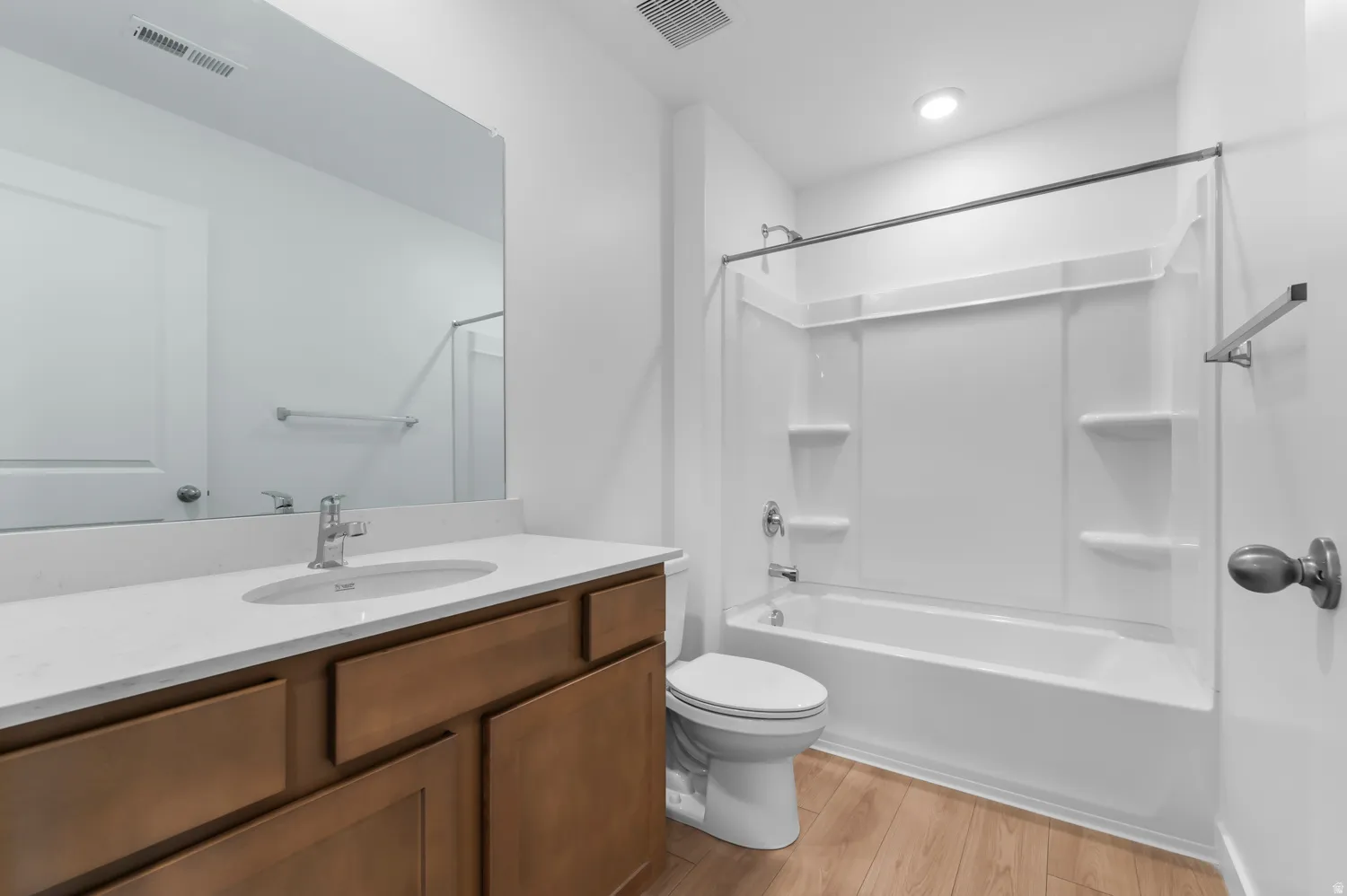 Bathroom featuring a white bathtub and shower combination with an integrated shelving system, a single vanity with a white countertop and wood-finish cabinetry, a large frameless mirror, and wood-finish flooring