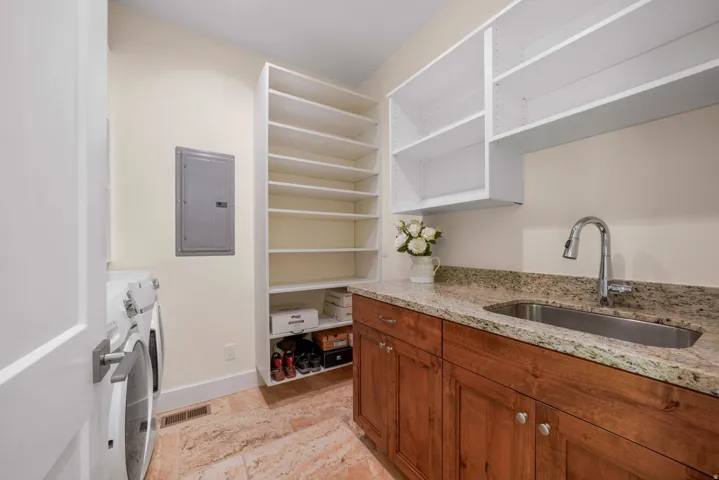 Laundry area featuring ample built-in shelving, stone tile flooring, a utility sink, and wood cabinetry with a granite countertop