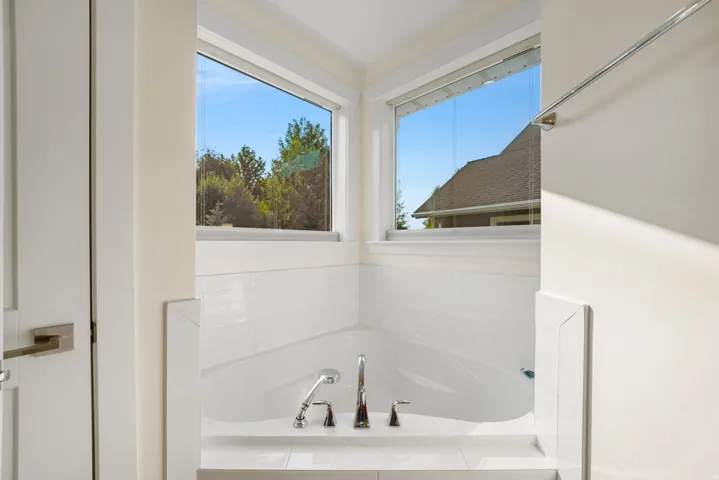 Corner soaking tub with polished chrome fixtures, white tile surround, and dual casement windows