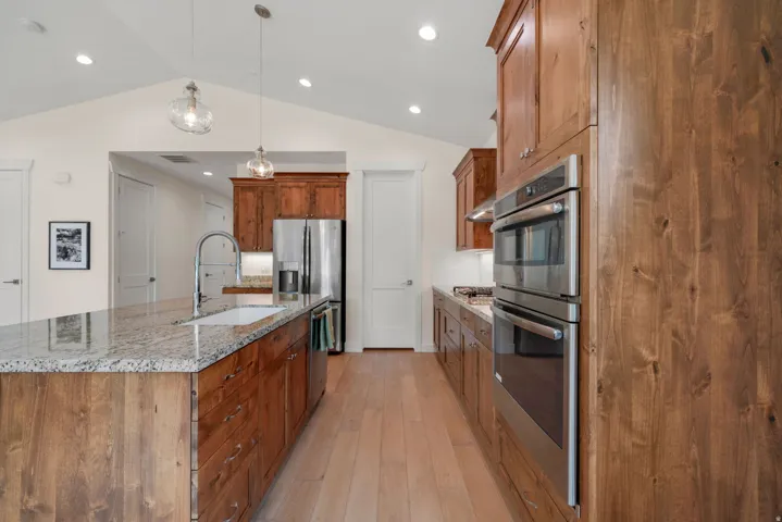Kitchen featuring a central island with a built-in sink and granite countertop, light wood-finish flooring, custom wood cabinetry, stainless steel double wall ovens, and recessed lighting
