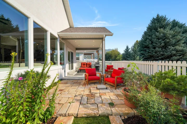 Outdoor stone patio featuring a covered porch, white picket fencing, and mature evergreen landscaping
