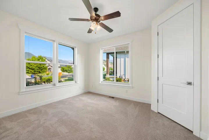 Bright and airy bedroom featuring abundant natural light from dual casement windows, neutral carpet flooring, ceiling fan with integrated lighting, and direct access to an ensuite or adjacent space via white panel door
