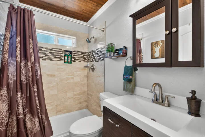 Bathroom featuring a wood-plank ceiling, tiled shower surround with accent mosaic, recessed shower niche, single vanity with integrated sink, and dark wood-finish medicine cabinet