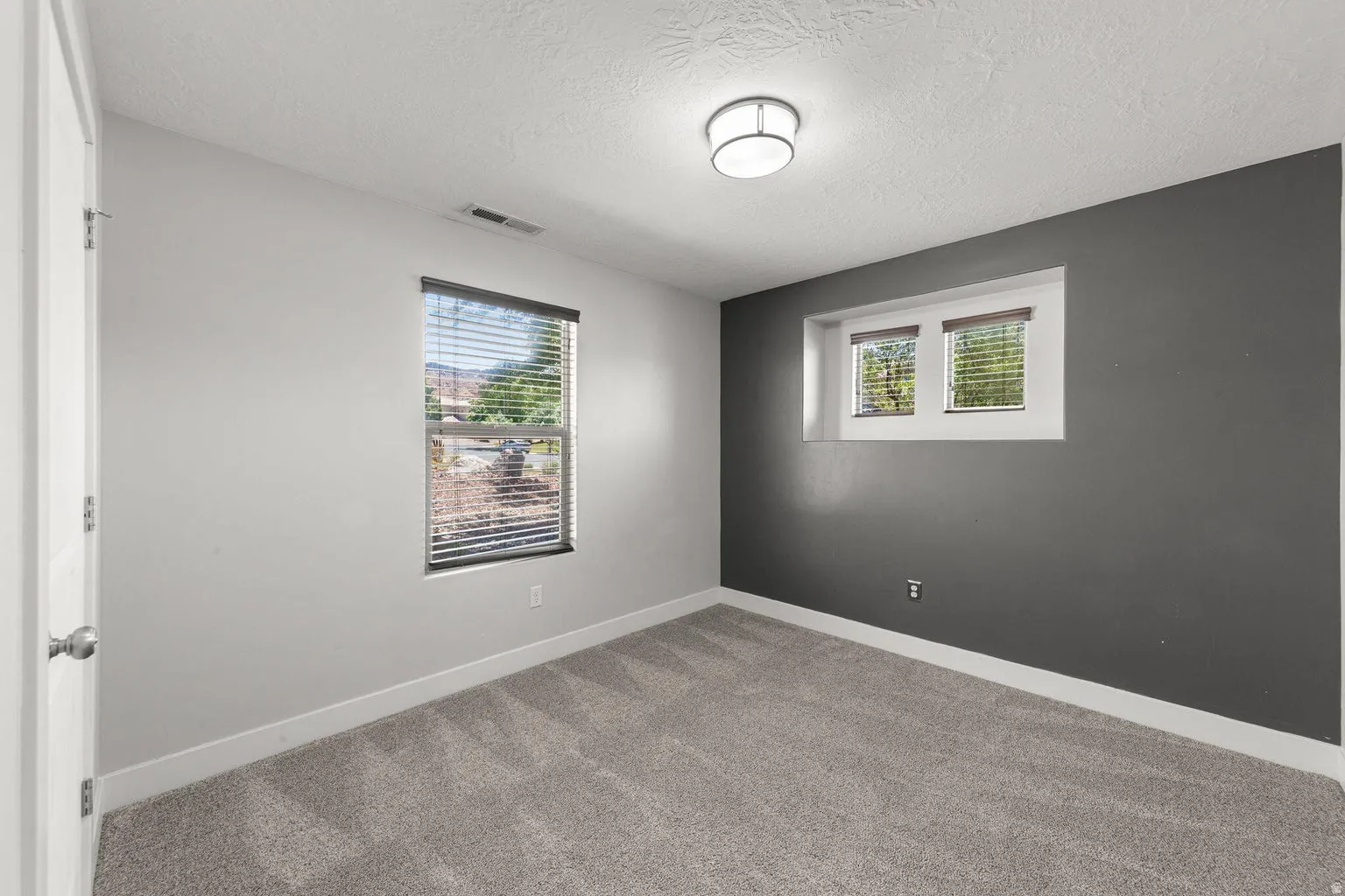 Interior room featuring light gray carpeting, a single window with horizontal blinds, and a contrasting dark gray accent wall