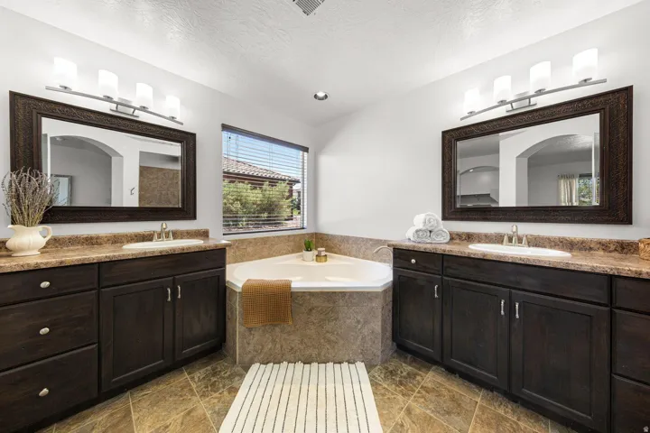 Bathroom featuring a corner soaking tub with tile surround, dual dark wood vanities with integrated sinks and stone-finish countertops, and large framed mirrors with contemporary lighting fixtures