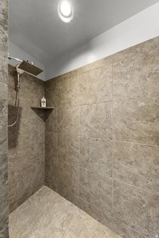 Walk-in shower with textured tile walls, coordinating tile flooring, a built-in corner shelf, and a ceiling-mounted rainfall showerhead