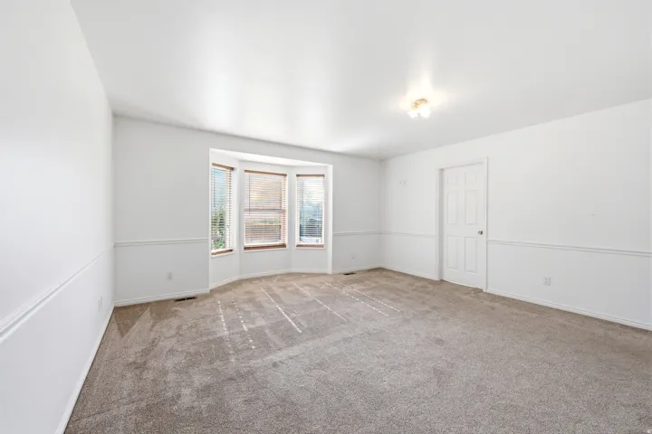 Bright room featuring a large bay window with wood-finish blinds, light-colored carpet flooring, white walls with chair rail molding, and a paneled door